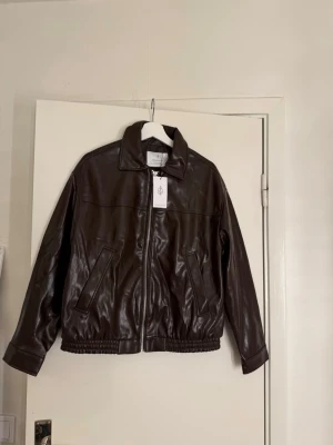 Brun skinnjacka från Stradivarius - Säljer en mörkbrun skinnjacka från Stradivarius med klassisk krage och dragkedja framtill. Jackan har två snedställda fickor, elastisk nederkant och långa ärmar med knappdetalj vid mudden. Perfekt för dig som vill ha en stilren och trendig look.