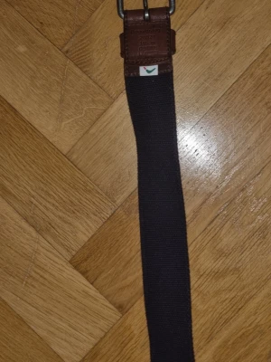 Mörkblå bälte från Boomerang - Snygg mörkblå bälte fån Boomerang med klassisk design. Bälte  har detaljer i brunt skinn vid spännet och är tillverkad i ett slitstarkt tygmaterial. Perfekt för dig som vill ha en stilren och enkel look.