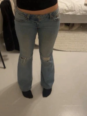 Ljusblå bootcut jeans med slitningar - Säljer ett par ljusblå jeans med bootcut passform och slitna detaljer över knäna. Byxorna har låg midja och klassisk femficksdesign. Perfekta för dig som gillar en avslappnad och trendig look.Storlek 38 men passar 36/38