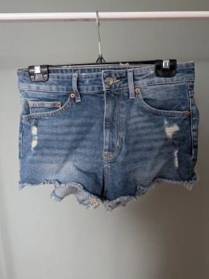 Blå jeansshorts med fransig kant - Säljer ett par klassiska blå jeansshorts med slitna detaljer och fransig kant nertill. Shortsen har fem fickor, bälteshällor och knappgylf. Perfekta för sommaren och ger en avslappnad vibe.