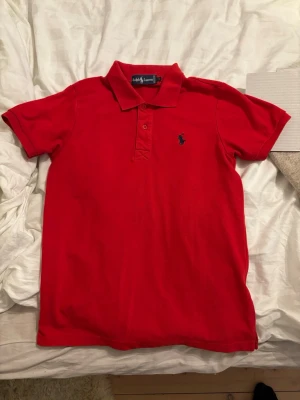 Röd pikétröja från Ralph Lauren - Klassisk röd pikétröja från Ralph Lauren med korta ärmar och krage. Tröjan har knappar framtill och den ikoniska broderade loggan i mörkblått på bröstet. Perfekt skick!