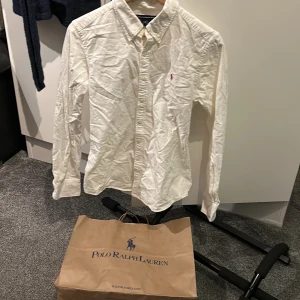 Vit skjorta från Polo Ralph Lauren - Klassisk vit skjorta från Polo Ralph Lauren med knappar framtill och broderad logga i rött på bröstet. Skjortan har lång ärm och krage, perfekt för en stilren look.storleken är 12 vilket motsvarar cirka storlek xs
