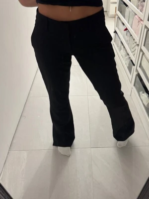 Svarta kostymbyxor med slits - Säljer ett par svarta kostymbyxor med raka ben och diskret slits vid benslutet. Byxorna har hög midja, bälteshällor och fickor på sidorna. Perfekta för en clean och stilren look. Materialet känns mjukt och följsamt. Säljer då de är för långa för mig och inte kommer till användning. 
