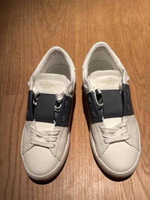 Valentino Garavani vita sneakers - Säljer ett par vita Valentino Garavani sneakers i skinn med bred mörkblå rem över vristen och coola nitar på hälen. Klassisk rund tå, vita snören och platt sula. Märkeslogga på plösen och sula. Perfekta för dig som gillar exklusiva och stilrena skor.