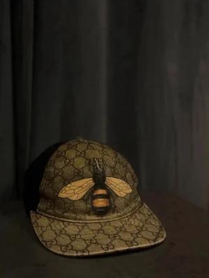 Gucci keps med bi och GG-mönster - Säljer en snygg Gucci keps med klassiskt GG-mönster i beige och brunt samt en stor broderad bi framtill. Baksidan är svart mesh med justerbart band i grönt och rött. Perfekt accessoar för dig som vill sticka ut med lyxig streetstyle.
