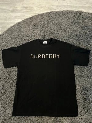 Svart Burberry t-shirt med logga - Säljer en svart t-shirt från Burberry med stor logga i beige och vit på bröstet. Klassisk rund hals och korta ärmar. Materialet är mjuk bomull som känns skönt mot huden. Perfekt för dig som gillar stilrena designer och vill ha ett statement-plagg. Storlek L och aldrig använd. Hör av dig vid funderingar.
