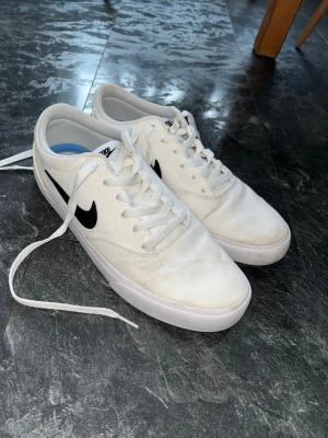 Vita Nike sneakers  - Säljer ett par klassiska vita Nike sneakers med svart swoosh på sidan. Skorna har snörning, rund tå och platt sula. Ovandelen är i textil och insidan har en blå innersula med Solesole-logga. Perfekta till vardagslooken! Använda endast ett fåtal gånger pga för liten storlek. 