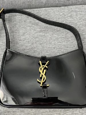 Svart axelväska från Saint Laurent - Säljer en svart axelväska från Saint Laurent i glansigt skinn med ikoniskt YSL-emblem i guld framtill. Väskan har en enkel, stilren form och justerbart axelband. Perfekt för dig som vill ha en lyxig och tidlös accessoar.