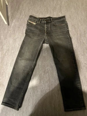 Svarta Diesel jeans straight fit - Säljer ett par svarta Diesel jeans med snyggt slitna detaljer och kontrastsömmar. Klassisk femficksmodell med rakt ben och diskret röd Diesel-logga på fickan. Jeansen är i ett robust denimtyg och har en tidlös look som passar till det mesta. Org pris 900kr men har använts hyfsat  mycket 