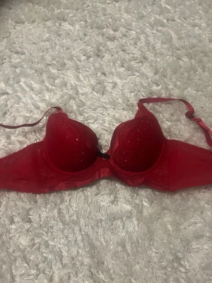 Röd spets-bh med strass från Hunkemöller - Snygg röd bh från Hunkemöller med spetsdetaljer och små strass-stenar på kuporna. Axelbanden är justerbara och bh:n har en liten rosett mellan kuporna. Perfekt för dig som vill ha något extra fint under kläderna.