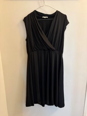 Svart omlottklänning från Rosebud - Säljer en svart omlottklänning från Rosebud i mjukt och följsamt material. Klänningen har v-ringad omlottdesign, markerad midja och är ärmlös. Perfekt för dig som gillar enkel men stilren look.