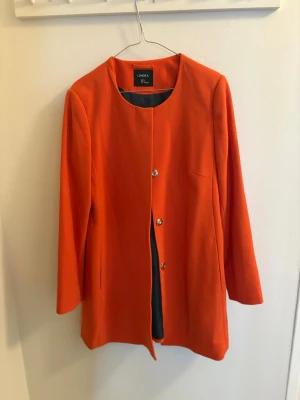 Orange kappa från Lindex, M - Säljer en snygg orange kappa från Lindex i storlek M. Jackan har raka linjer, rund halsringning och stängs med tre silverfärgade knappar framtill. Lätt fodrad och i ett mjukt tyg som känns bekvämt. Perfekt för dig som vill sticka ut med färg.