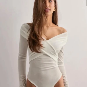 Vit omlott offshoulder topp från Nelly  - Snygg vit omlott topp med offshoulder-design och långa ärmar. Toppens material är tunt och stretchigt, vilket ger en smickrande passform. Perfekt för dig som vill ha en trendig och stilren look.