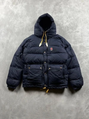 Mörkblå dunjacka från Fjällräven - Säljer en mörkblå dunjacka från Fjällräven med huva och gula snören. Jackan har två stora fickor framtill med knappar och klassisk Fjällräven-logga på bröstet. Perfekt för kalla dagar och riktigt skön att bära. Passformen är normal och jackan är vadderad för extra värme.