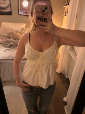 Vit linnetopp med knappar - Säljer en söt vit linnetopp med smala axelband och knappar framtill. Toppen har en lätt rynkad byst och markerad midja som ger en feminin siluett. Perfekt till jeans eller kjol för en chill sommarlook.