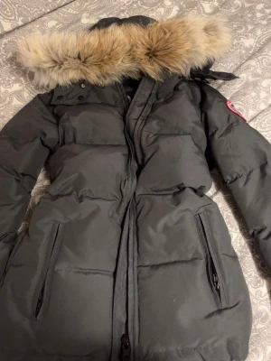 Mörkgrå Canada Goose jacka - Säljer en mörkgrå dunjacka från Canada Goose med avtagbar huva och snygg pälsdetalj. Jackan har långa ärmar, två stora fickor med dragkedja och klassisk patch på ärmen. Perfekt för kalla vinterdagar och riktigt varm. Såklart är pälsen äkta