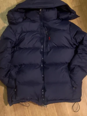 Mörkblå pufferjacka från Ralph Lauren - Säljer en mörkblå pufferjacka från Polo Ralph Lauren med röd broderad logga på bröstet. Perfekt för kalla dagar och passar till i princip allting.