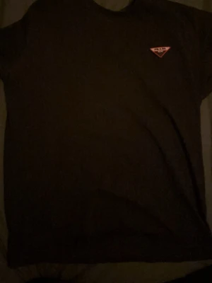 Svart t-shirt från Prada - Svart t-shirt från Prada med klassisk rund hals och korta ärmar. På bröstet sitter den ikoniska röda Prada-loggan i trekant. T-shirten är i mjuk bomull och har en clean, stilren look som funkar till allt.