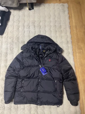 Svart pufferjacka storlek M-L, från Polo Ralph Lauren - Svart pufferjacka från Polo Ralph Lauren med röd broderad logga på bröstet. Jackan har huva, två sidofickor med dragkedja och RL-detaljer på zip. Materialet är polyester och bomull, vilket gör den både lätt och varm. Perfekt för kalla dagar.