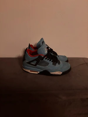 Nike Air Jordan 4 Travis Scott blå - Säljer ett par Nike Air Jordan 4 Travis Scott i blå mocka med svarta och röda detaljer. Skorna har svart snörning, meshpaneler på sidorna och specklade svarta hälpaneler med Jumpman-logga och Cactus Jack-tryck. Röd insida och vit sula med svart och röd detalj.