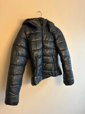 Blå/grön pufferjacka med huva - Säljer en jacka med huva och dragkedja framtill. Jackan är quiltad och har två fickor med dragkedja. Perfekt för kalla dagar när du vill hålla dig varm och ändå se snygg ut.