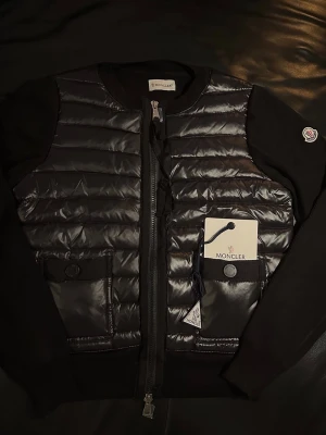 Moncler Cardigan  - Snygg svart pufferjacka från Moncler med quiltad framsida och två fickor med knappstängning. Jackan har dragkedja och Moncler-logga på ärmen. Materialmix med polyester och stickade detaljer på ärmar och sidor. Perfekt för dig som gillar exklusiv streetstyle.