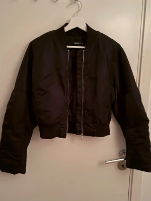 Svart bomberjacka från Gina Tricot - Säljer en svart bomberjacka från Gina Tricot. Jackan har en croppad passform, ribbade muddar och dragkedja framtill. Tillverkad i ett glansigt syntetmaterial som ger en cool och modern look. Perfekt till alla dina outfits! Helt oanvänd!!