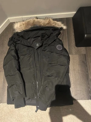 Svart dunjacka från Canada Goose - Säljer en svart dunjacka från Canada Goose med avtagbar huva och snygg fuskpäls. Jackan har ribbade muddar, dragkedja och knappar framtill samt klassisk patch på ärmen. Perfekt för kalla vinterdagar och riktigt varm.