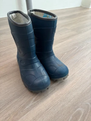 Mörkblå fodrade stövlar - Säljer ett par mörkblå stövlar med grov sula och mjukt, ljust foder på insidan. Skorna har en robust design med mönstrad yta och är perfekta för kalla och blöta dagar. Tillverkade i syntetmaterial och håller fötterna varma och torra.