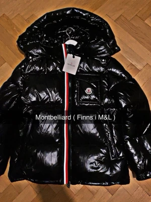 Svart pufferjacka från Moncler - Säljer en svart glansig pufferjacka från Moncler med huva och ikonisk logga på bröstet. Jackan har en rödvit rand längs dragkedjan och praktiska fickor. Perfekt för kalla dagar och riktigt snygg streetstil.