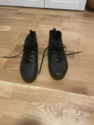 Svarta Nike fotbollsskor med snörning - Säljer ett par svarta Nike phantom fotbollsskor med högre skaft och snörning. Skorna har rund tå, platt sula med dobbar och en diskret Nike-logga på sidan. Tillverkade i syntetmaterial och designade för bra grepp på planen.