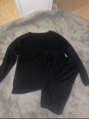 Svarta mjukisbyxor och tröja i storlek L - Säljer ett svart set i storlek L. Byxorna har resår i midjan och är gjorda i ett mjukt och bekvämt material, perfekta för chill dagar hemma. Klassisk rak passform och enkel design utan detaljer. Aldrig använt. Vet inte vad det är för märke men köpte dom för 499kr