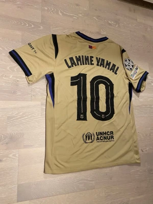 Beige Barcelona 25/26 T-shirt Lamine Yamal  - •Hej! Säljer en jätte snygg Beige Barcelona 25/26 fotbolls T-shirt med Lamine Yamal på ryggen ⚽️👕 •Den är inte använd bara testad.  •Det är storlek M och den har inga hål eller liknande. •Måtten är på dem sista bilderna 📐.  •Priset kan diskuteras och skickar inom 24 Tim😊💨