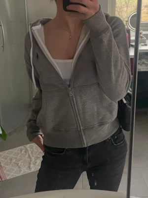 Grå hoodie från Polo Ralph Lauren - Grå zip-hoodie från Polo Ralph Lauren med vit dragsko i huvan och klassisk blå logga på bröstet. Mjuk bomullskänsla, fickor framtill och ribbade muddar.