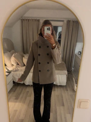 Beige skepparkavaj med hög krage - Stilren beige skepparkavaj med dubbelknäppning och hög krage. Jackan har en klassisk passform och svarta knappar framtill. Perfekt för dig som vill ha en tidlös look med snygga detaljer. Den är liten i storleken då jag egentligen har XS!!🤍