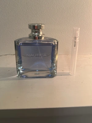 Nautica Voyage parfym blå flaska - Fräsch parfym från Nautica i en stilren, rektangulär glasflaska med blått innehåll och silverfärgad kork. Medföljer även ett litet provrör. Perfekt för dig som gillar marina och fräscha dofter.