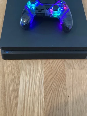 PlayStation 4 Slim med LED-kontroll - Svart PlayStation 4 Slim med en transparent spelkontroll som har färgglada LED-lampor. Konsolen har en stilren, rektangulär design och kontrollen är gjord av plast med synliga komponenter. Perfekt för gamingkvällar och långa spelsessioner. finns att hämta i skåne mvh säljaren!