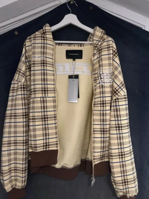 Rutig hoodie från Coldculture - Snygg hoodie från Coldculture i beige och brunt rutigt mönster med blå detaljer. Hoodien har dragkedja, bruna muddar och huva. På bröstet finns en broderad patch och logga. Perfekt för dig som gillar streetwear och vill sticka ut. Originalpris : 1200 kr