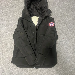 Svart dunjacka från Canada Goose - Snygg svart dunjacka från Canada Goose med huva och klassisk logopatch på ärmen. Jackan har en quiltad design, dragkedja framtill och är perfekt för kalla dagar. Passar dig som vill ha både stil och värme under vintern.
