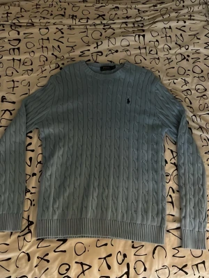 Ljusblå kabelstickad tröja Polo Ralph Lauren - Ljusblå kabelstickad tröja från Polo Ralph Lauren med rund halsringning och broderad logga på bröstet. Tröjan har ribbade muddar vid ärmslut och nederkant, perfekt för en stilren look.