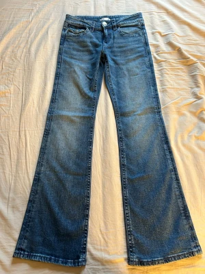 Blå bootcut jeans från Gina Tricot - Snygga blå bootcut jeans från Gina Tricot i klassisk denim med fem fickor och knappgylf. Jeansen har en utsvängd passform från knät och är tillverkade i bomullsmaterial. Perfekta för dig som gillar en retroinspirerad look.