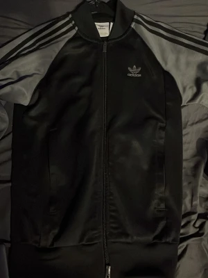 Hoodie   - Klassisk svart zip hoodie från Adidas med grå detaljer och de ikoniska tre ränderna på ärmarna. Hoodien  har dragkedja framtill, lång ärm och Adidas-logga på bröstet. Perfekt för en sportig och avslappnad stil.