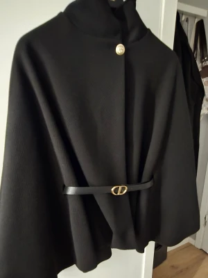 Svart cape-kappa med bälte - Stilren svart cape-kappa med hög krage och elegant bälte i midjan med gulddetalj. Kappan har en knapp framtill och ett svepande, vid siluett som ger en cool och modern look. Perfekt för dig som vill sticka ut med en unik ytterjacka.