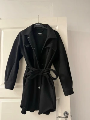 Svart overshirt från ONLY med bälte - Stilren svart kappa från ONLY med klassisk krage, knappar framtill och två bröstfickor. Jackan har ett matchande bälte i midjan för en snygg siluett och långa ärmar med knappdetaljer vid ärmslut. Perfekt för lager på lager och enkel att styla.