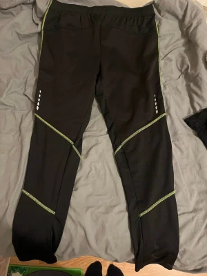 Svarta träningsbyxor från Crivit Sports - Svarta träningsbyxor från Crivit Sports med gröna sömmar och reflexdetaljer på benen. Byxorna har elastisk midja med snörning och dragkedjor vid bensluten för extra komfort. Perfekta för löpning eller gymmet.