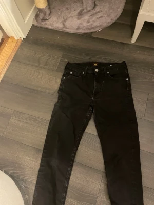Svarta Lee Austin jeans W29 L32 - Svarta jeans från Lee, modell Austin, med klassisk femficksdesign och raka ben. Jeansen har normal passform och är tillverkade i bomull med en snygg svart tvätt. Perfekta för en stilren och enkel look. Inte använda! 