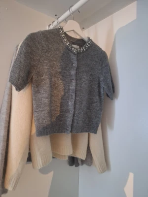 Grå kortärmad kofta med pärlkrage - Grå stickad kofta med korta ärmar och rund halsringning dekorerad med pärlor och stenar. Knäpps framtill med små knappar och har ribbade muddar vid ärmslut och nederkant. Perfekt för att lyfta en enkel outfit.