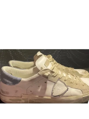Vita sneakers med beige mockadetaljer - Snygga vita sneakers från Philippe Model med beige mockadetaljer och ljusblå häl. Klassisk låg modell med snörning och rund tå. Skorna har en platt sula och ikonisk sköldlogga på sidan. Perfekta för en avslappnad streetstyle-look.