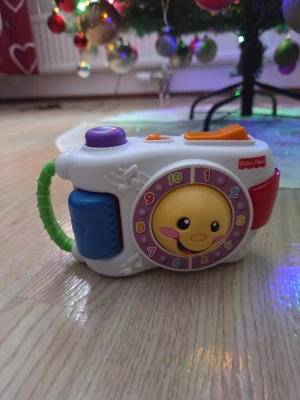 Fisher-Price leksakskamera - Rolig leksakskamera från Fisher-Price i vitt med färgglada detaljer som lila, orange, blå och grön. Kameran har en snurrande urtavla med siffror och en glad smiley, samt musiknoter på sidan. Perfekt för yngre barn som gillar att leka och utforska.