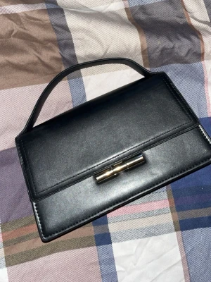 Svart kuvertväska i skinn med metallspänne  - Stilren svart kuvertväska i skinn med ett elegant metallspänne framtill. Väskan har en enkel, rektangulär form och ett kort handtag upptill. Perfekt för dig som gillar minimalistisk och klassisk design.